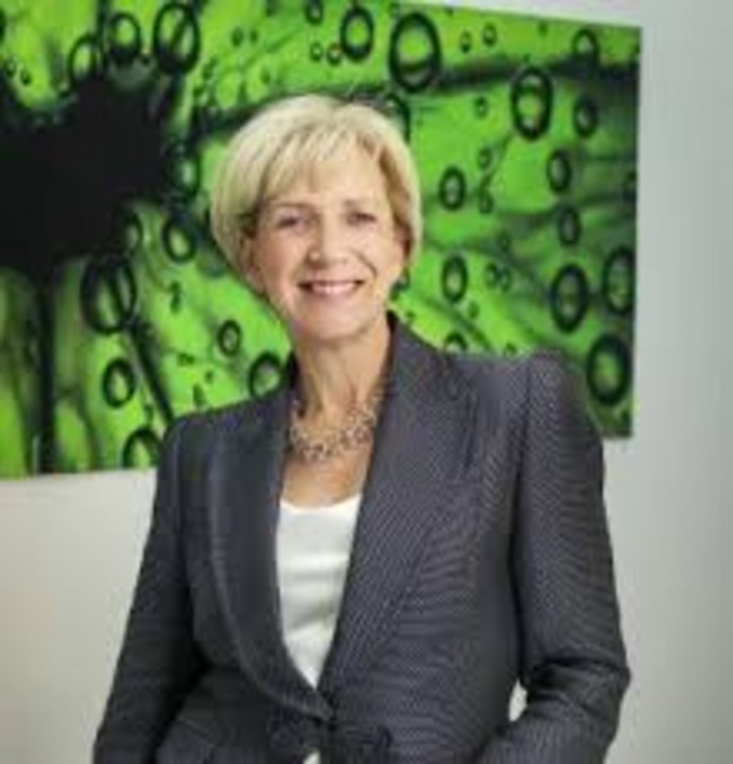 Smiling professional woman with short blonde hair wearing gray blazer and gold necklace, standing before vibrant green abstra
