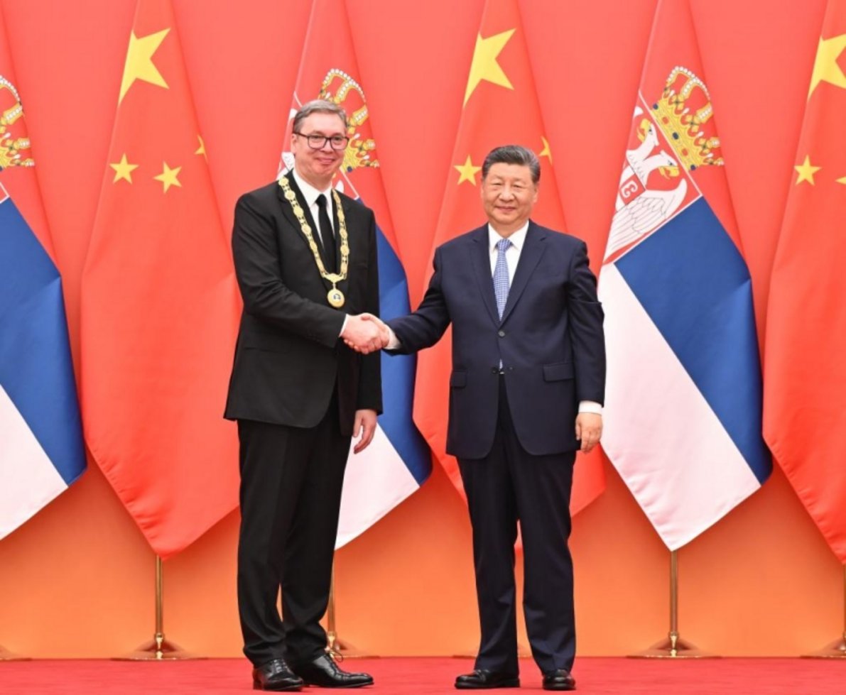 Two male leaders shaking hands in front of alternating Serbian and Chinese flags, one wearing ceremonial gold chain medal