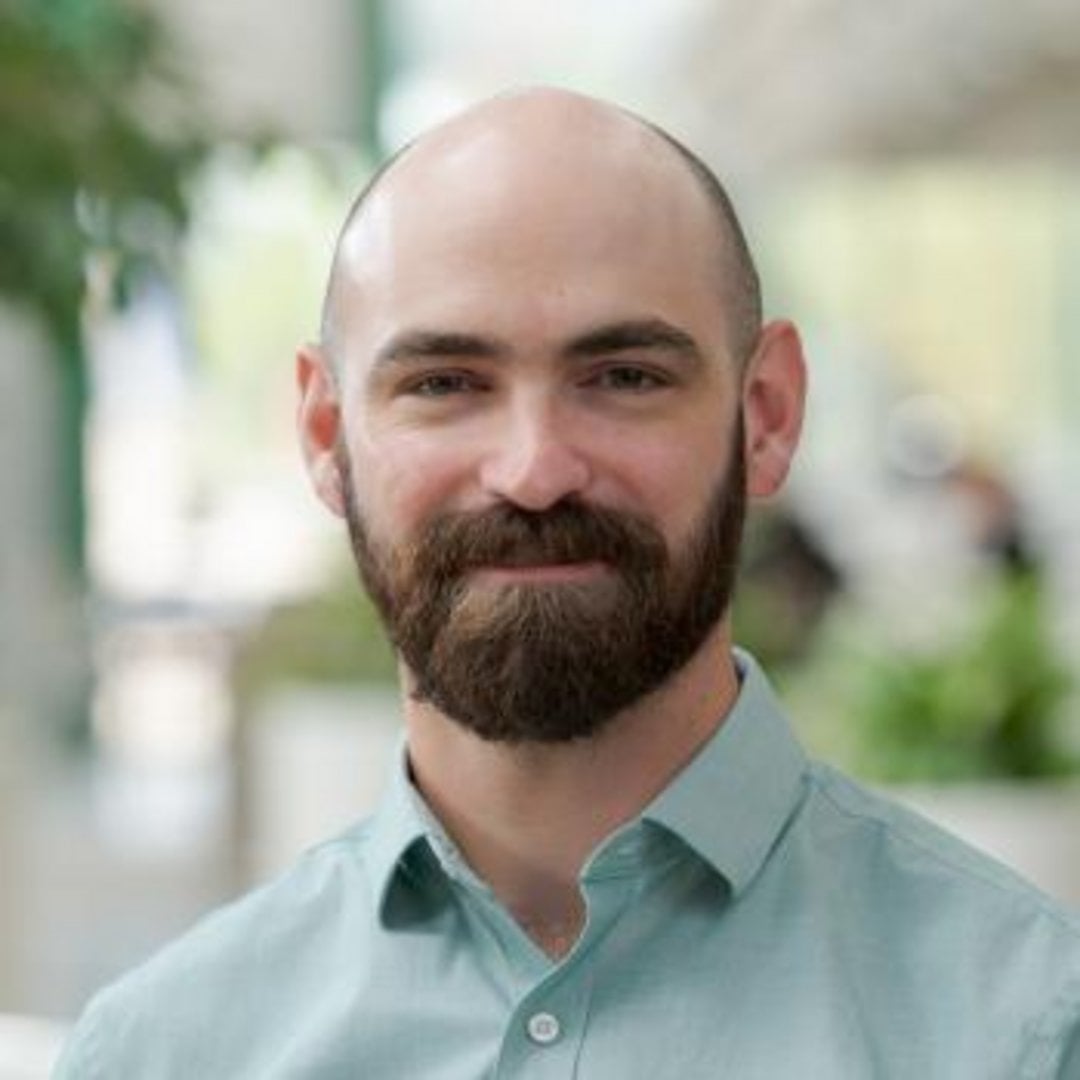 Bald man with a full dark brown beard wearing a light teal button-up shirt, smiling softly in an outdoor setting