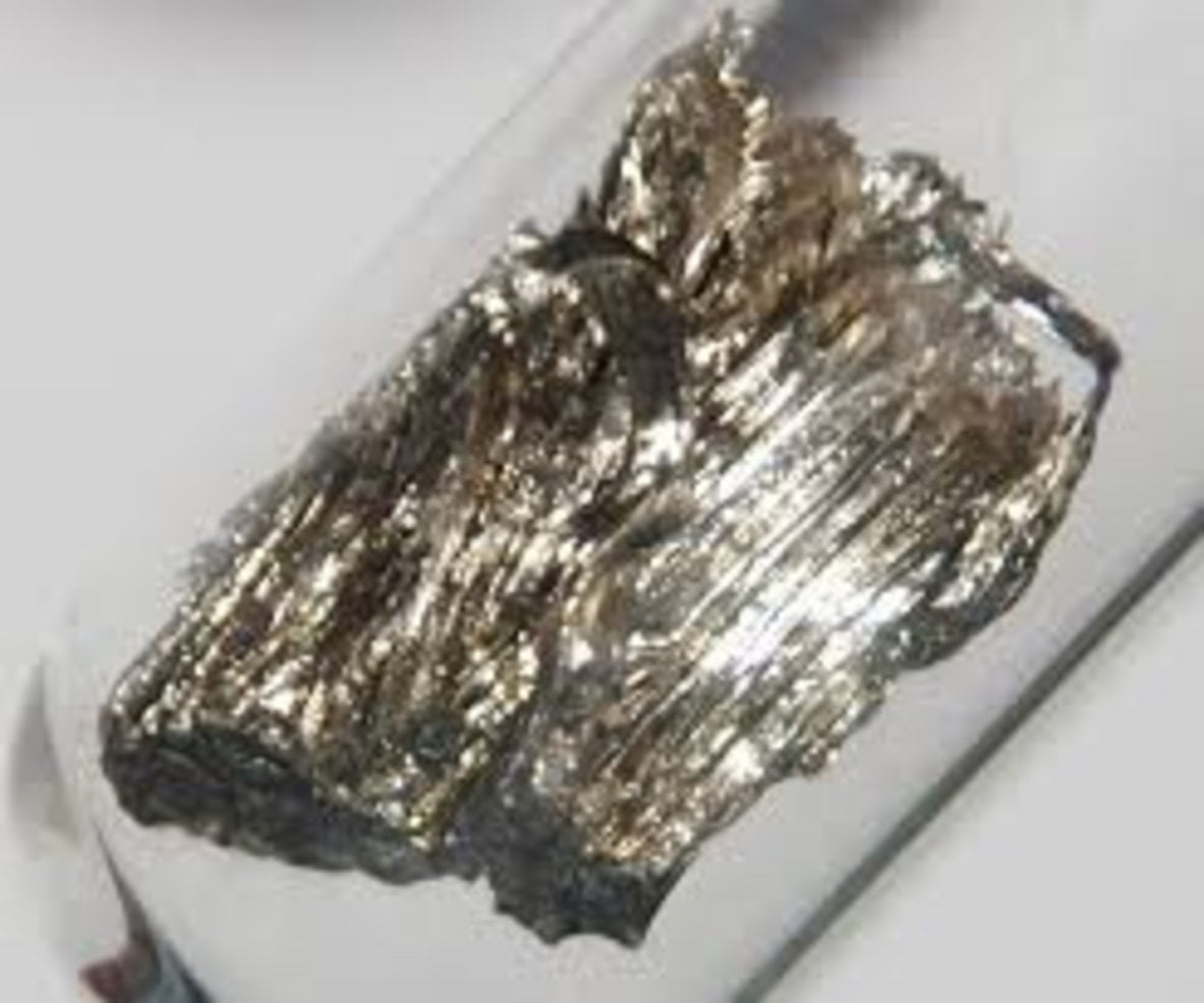 A crystalline chunk of dysprosium rare earth metal with silvery striated texture stored in a sealed borosilicate glass ampoul