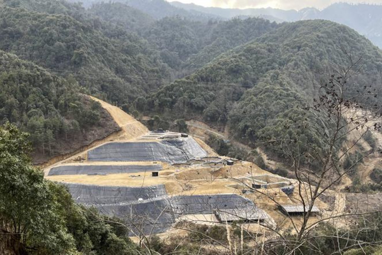 Engineered terraced landfill with black HDPE geomembrane liner panels on compacted earthen slopes surrounded by forested moun