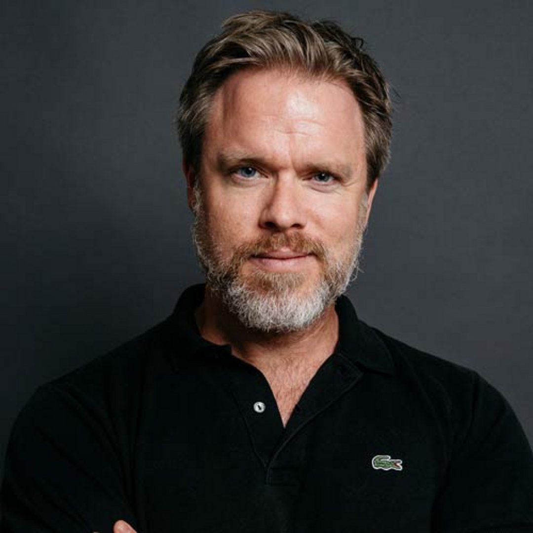 A middle-aged man with salt-and-pepper beard and blue eyes wearing a black Lacoste polo shirt, arms crossed, against gray bac