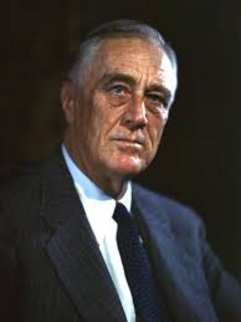 An older gentleman with silver hair wearing a dark suit jacket, white dress shirt, and navy polka dot tie against a dark back