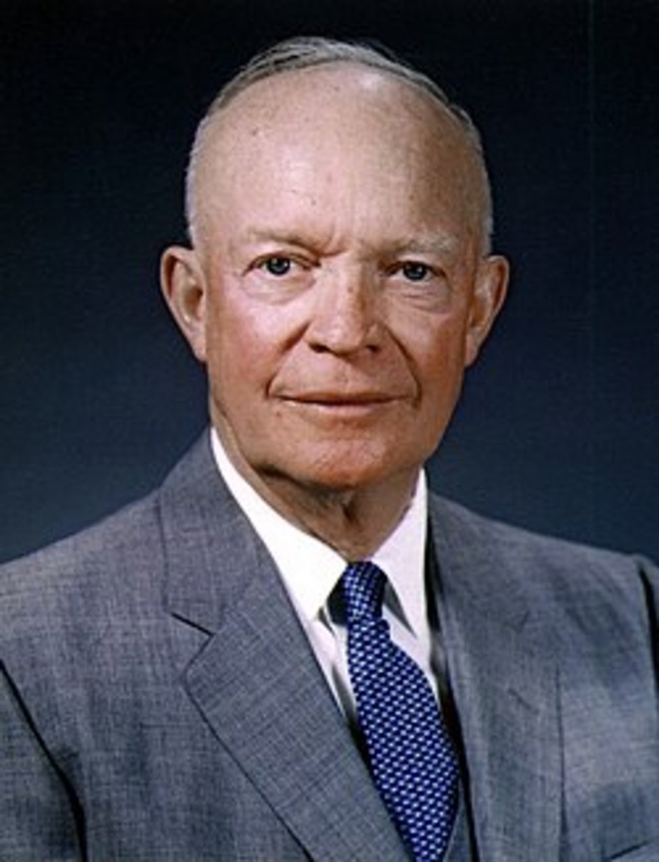 Older bald man with short white hair wearing a gray suit and blue patterned tie against a dark blue studio background
