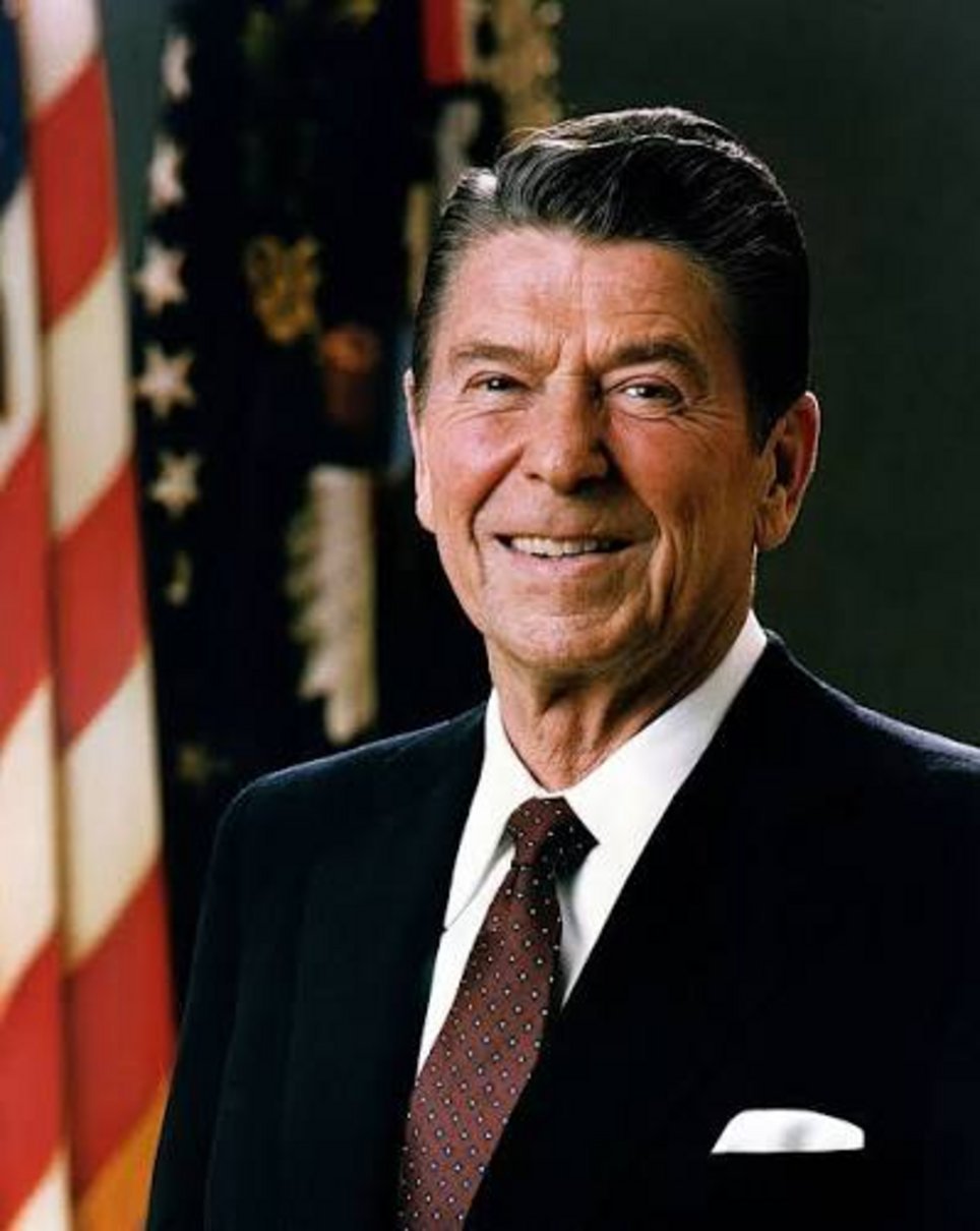 Older man with dark hair wearing black suit white shirt burgundy tie smiling in front of American flag and gold emblem