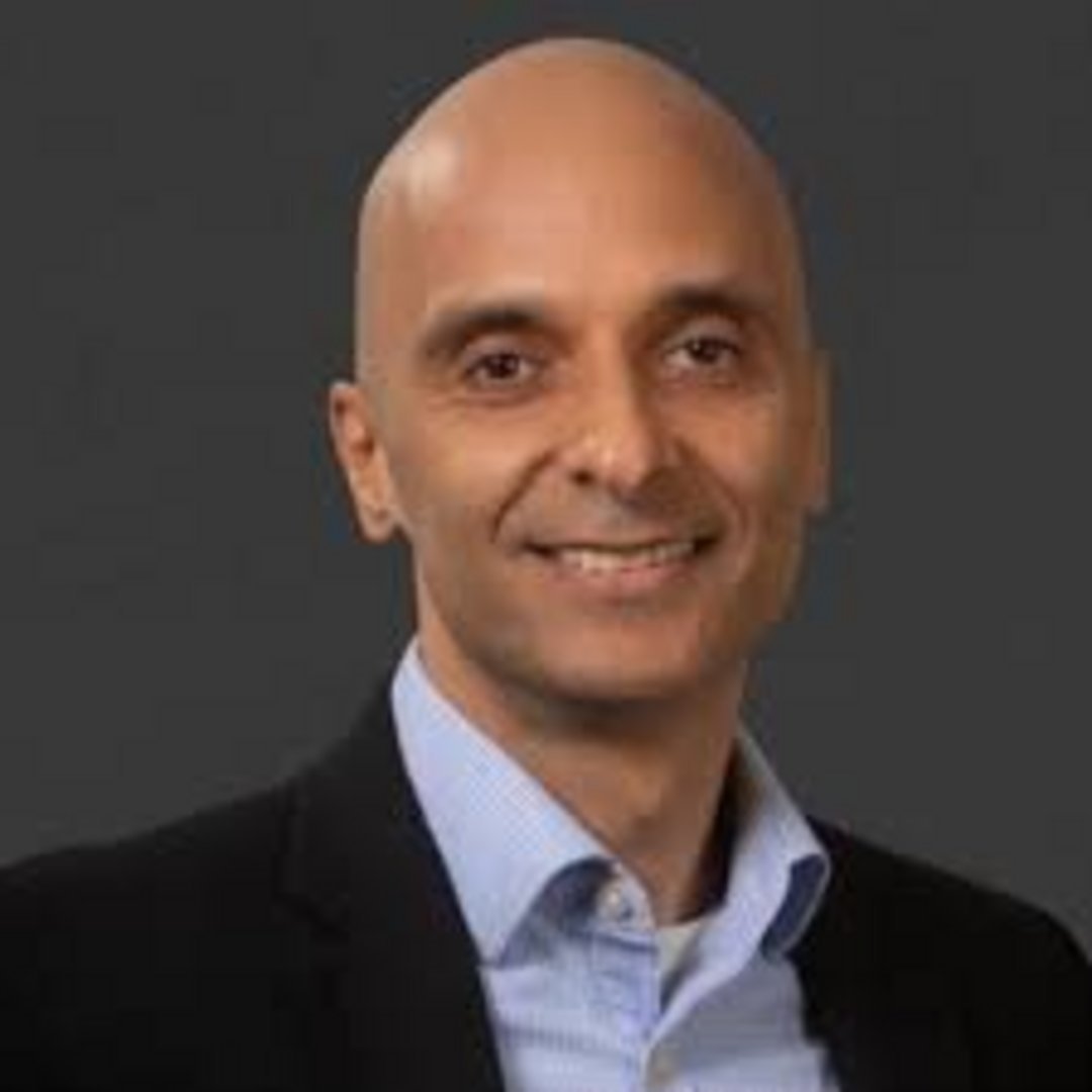 Middle-aged bald man with warm smile wearing black blazer over light blue collared shirt against neutral gray background