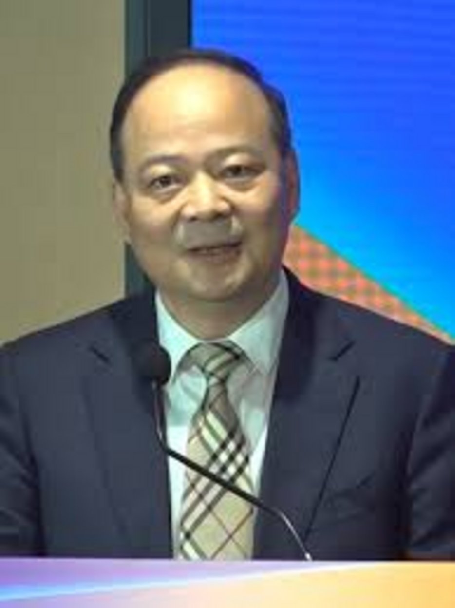 Middle-aged Asian man in navy blue suit and plaid Burberry tie speaking at a podium with a microphone in front of a blue scre