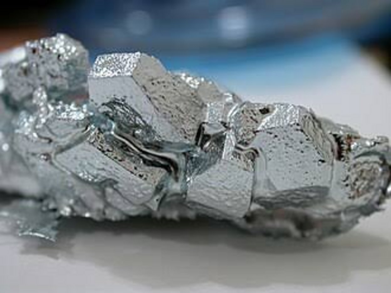 Crystalline gallium metal sample with faceted cubic formations and silvery liquid sheen at room temperature on white surface