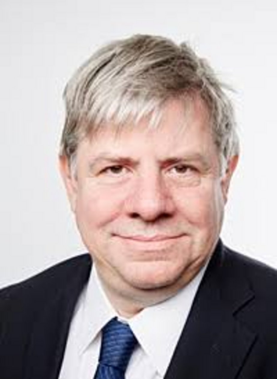 A middle-aged man with gray hair wearing a dark suit jacket, white dress shirt, and navy blue striped tie against a white bac