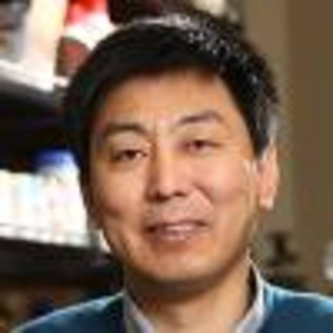 Middle-aged East Asian man with dark hair, subtle smile, wearing a blue sweater over collared shirt, standing near a bookshel