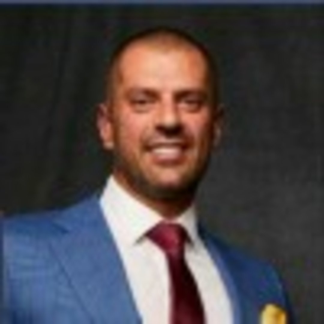 Smiling man in blue blazer, white dress shirt, burgundy tie, and gold pocket square against dark gray background
