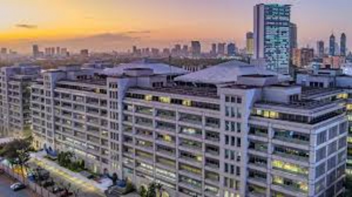 Large multi-story commercial office complex with illuminated windows at dusk, city skyline with high-rise towers visible in o