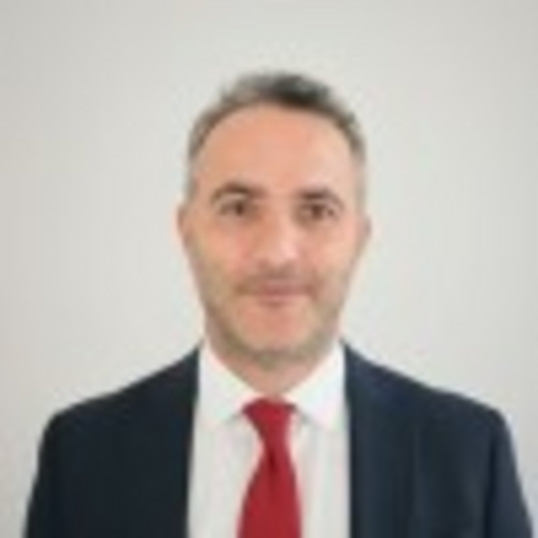 Middle-aged man with salt-and-pepper hair wearing a navy blue blazer, red tie, and white dress shirt against a gray backgroun
