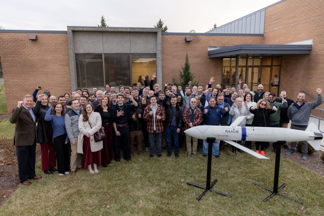 Large group of CoAspire aerospace engineers and staff celebrating outdoors beside a RAACM missile model display on a brick bu