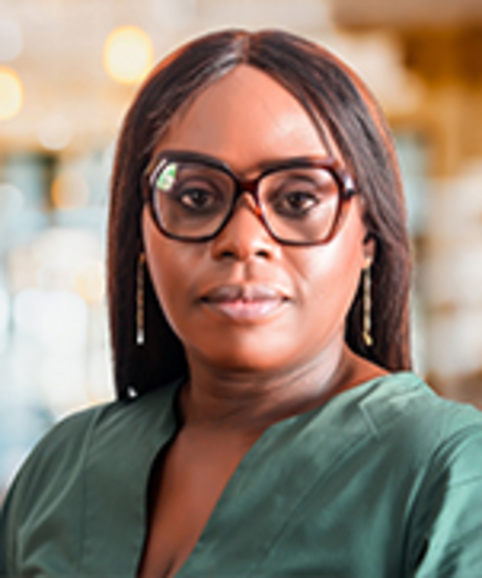 A professional Black woman with straight dark auburn hair, tortoiseshell glasses, gold drop earrings, and a teal green V-neck