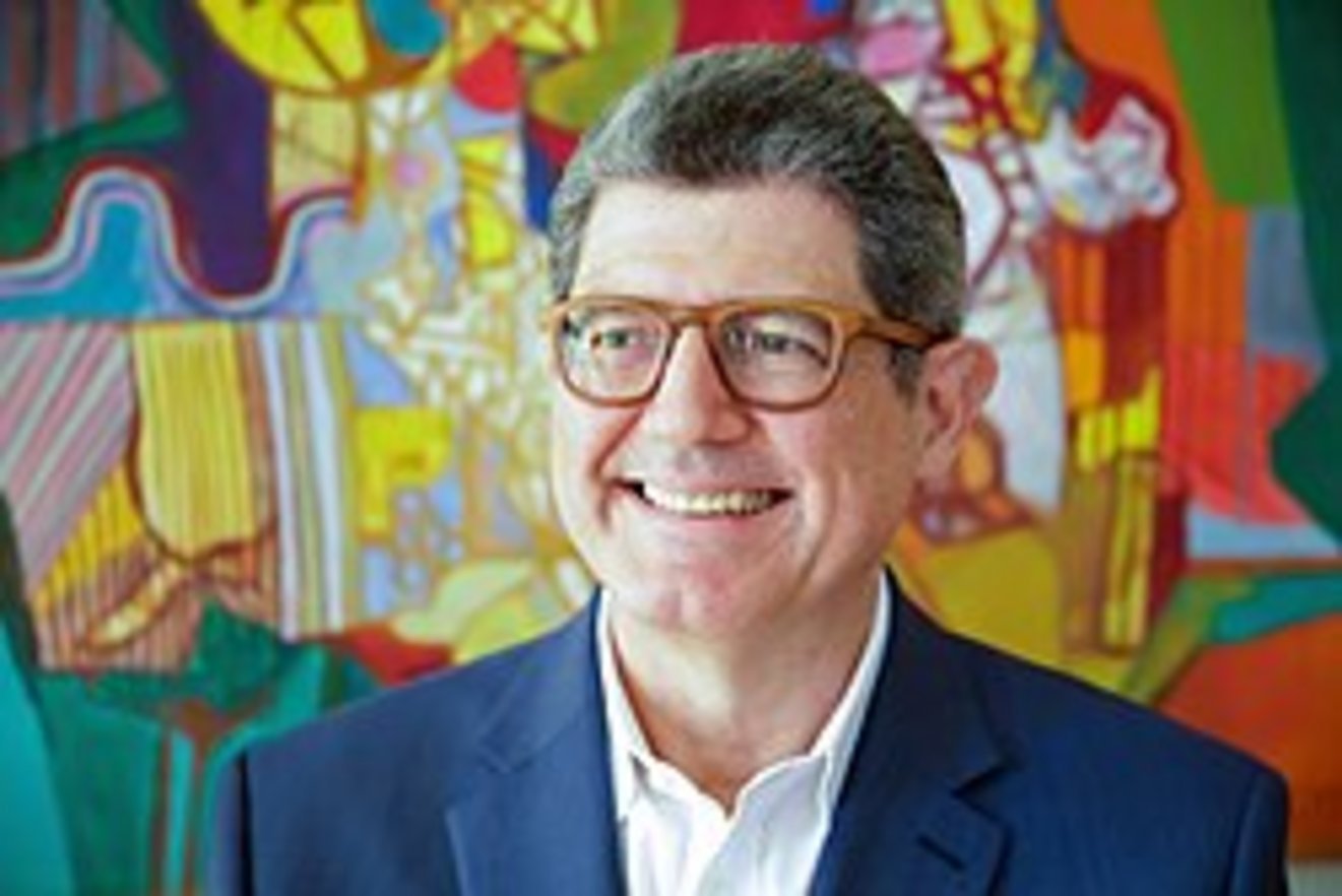 A smiling middle-aged man with short gray hair, brown rectangular glasses, and a navy blue blazer in front of a vibrant color