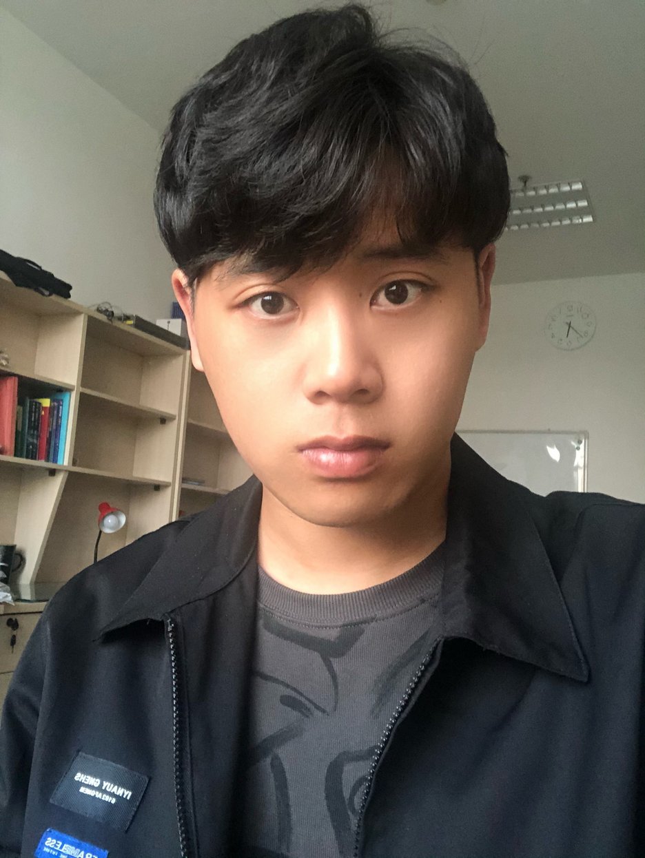 Young Asian man with black bowl-cut hair wearing a dark navy zip-up jacket over a gray graphic t-shirt, standing in a study r