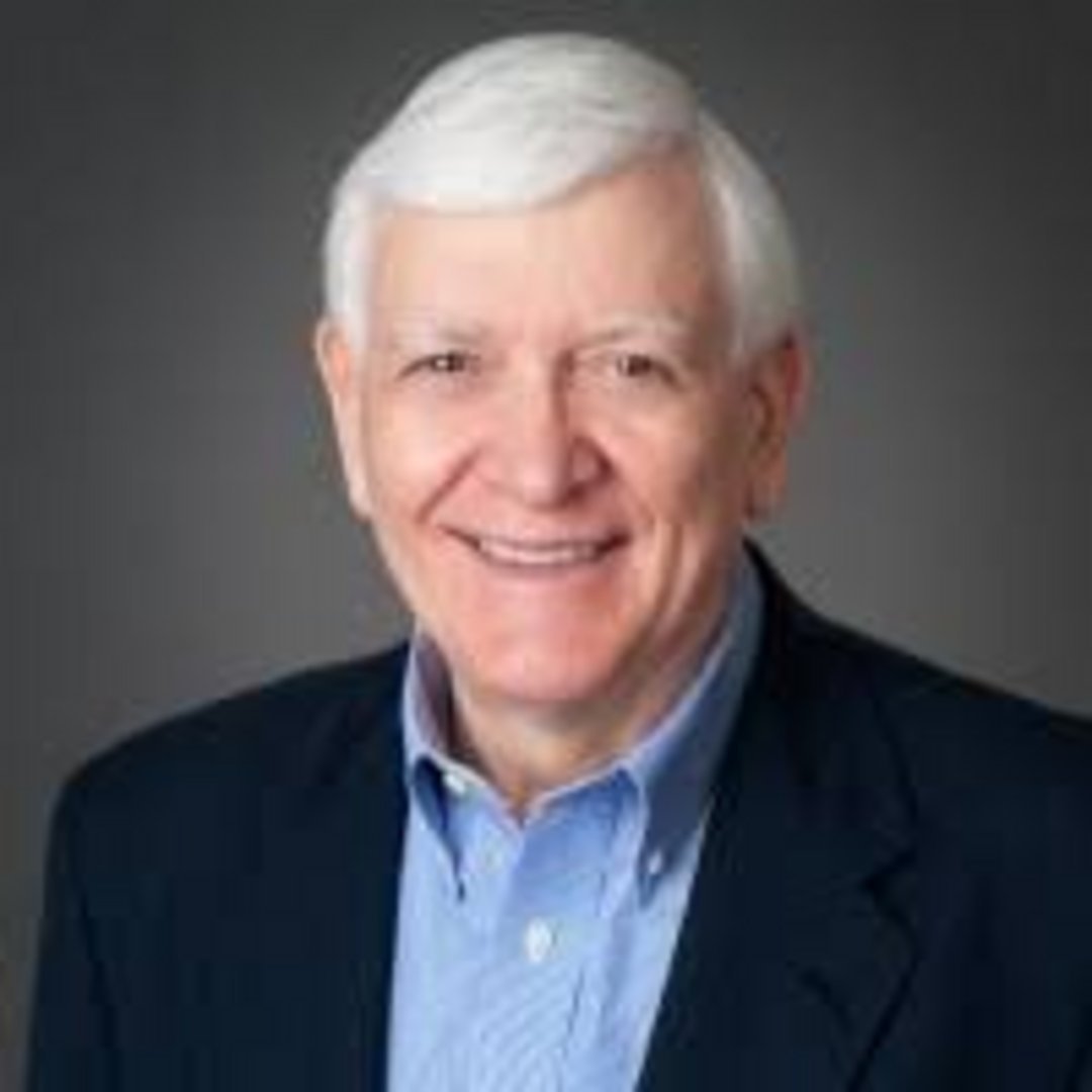 Older gentleman with white hair and warm smile wearing navy blazer and light blue button-down shirt against gray background