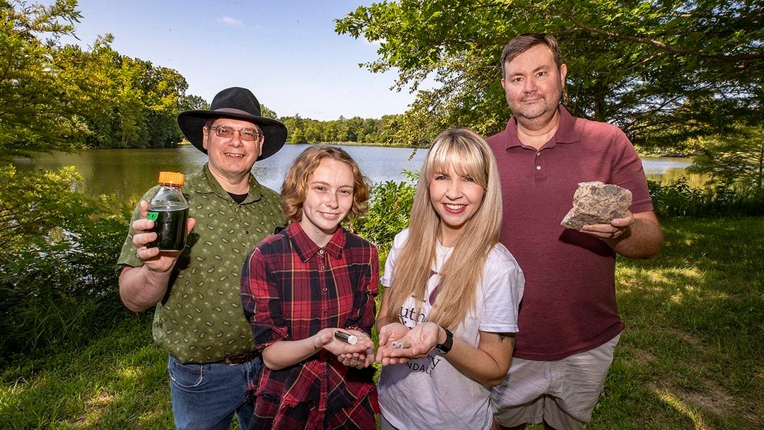 Four researchers pose near a tree-lined lake, holding a dark water sample bottle, small vials, and a rock specimen during an