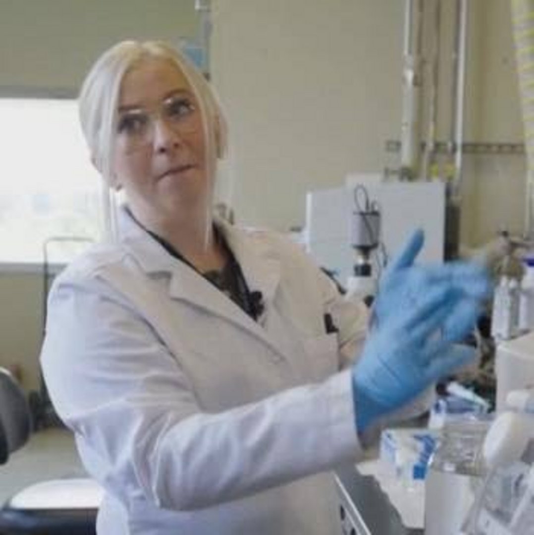 A blonde female scientist wearing a white lab coat and blue nitrile gloves working with equipment in a research laboratory
