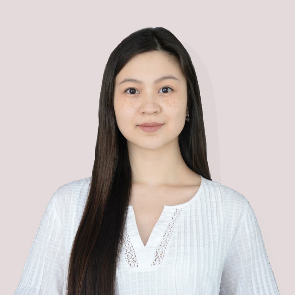 Young Asian woman with long straight dark hair and freckles wearing a white textured lace-trim blouse against a soft pink bac