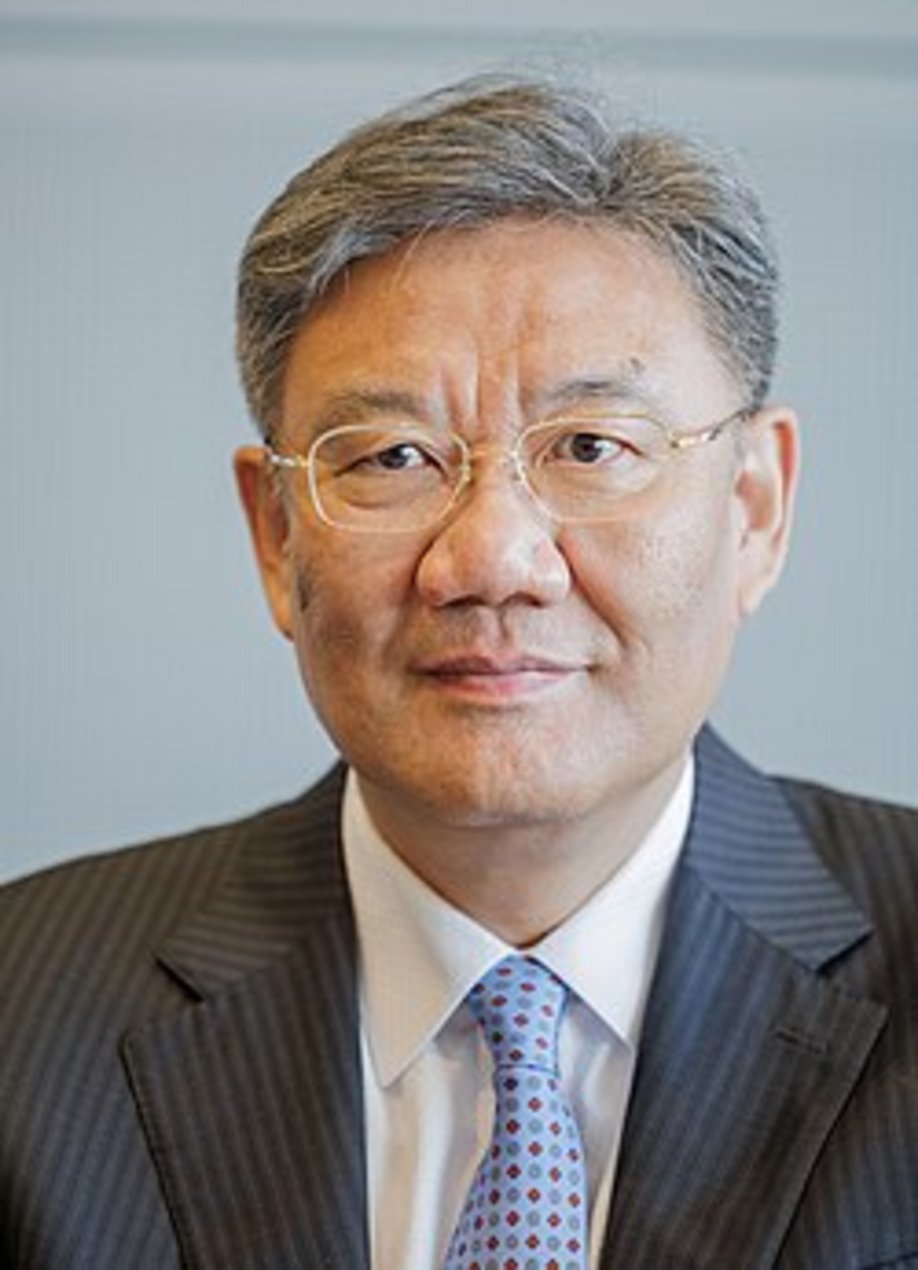 Older East Asian man with salt-and-pepper hair, gold-rimmed glasses, dark pinstripe suit, white shirt, and light blue pattern