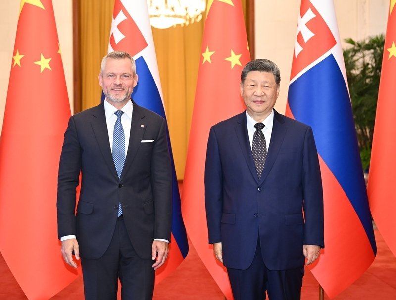 Two male diplomats in dark suits standing before alternating Chinese and Slovak flags in an ornate official reception hall