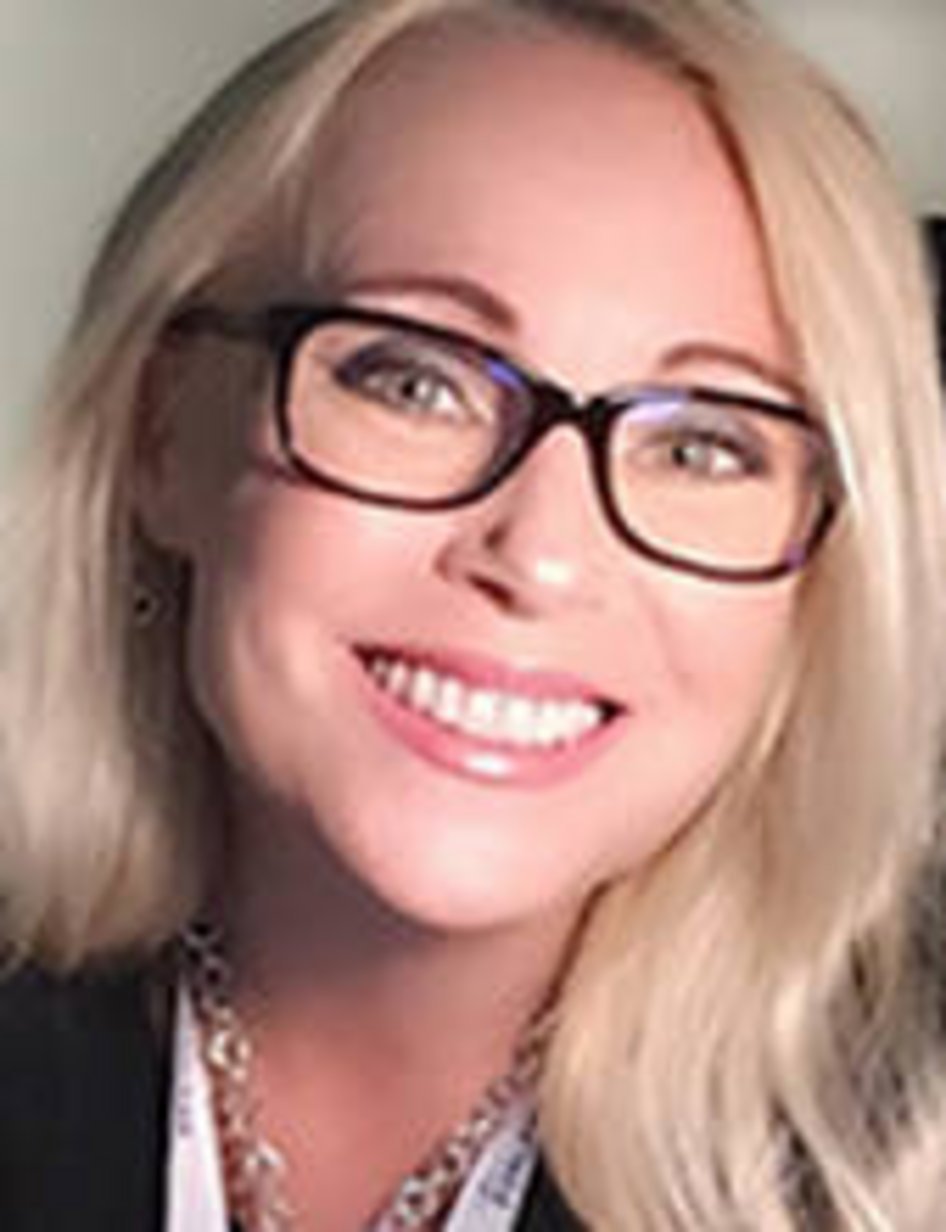 A smiling blonde woman wearing dark-framed rectangular glasses and a pearl necklace in a professional portrait setting