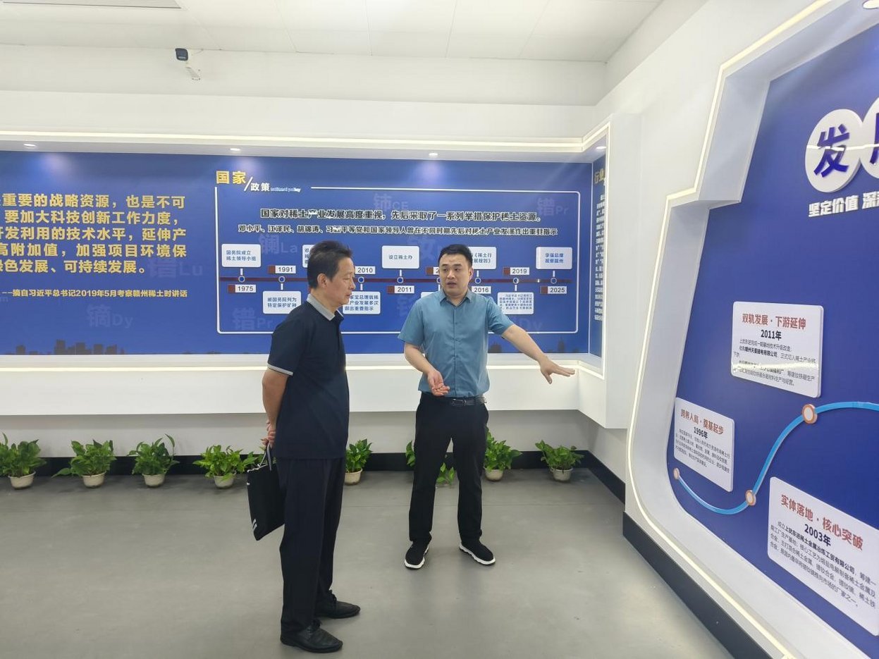 Two Chinese businessmen reviewing rare earth industry development timeline display panels in a blue-themed corporate exhibiti