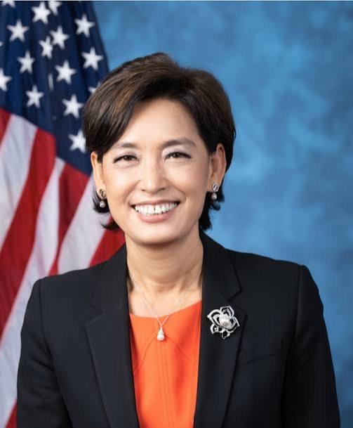 Woman in black blazer and orange top with pearl necklace and brooch smiling before American flag in official portrait setting