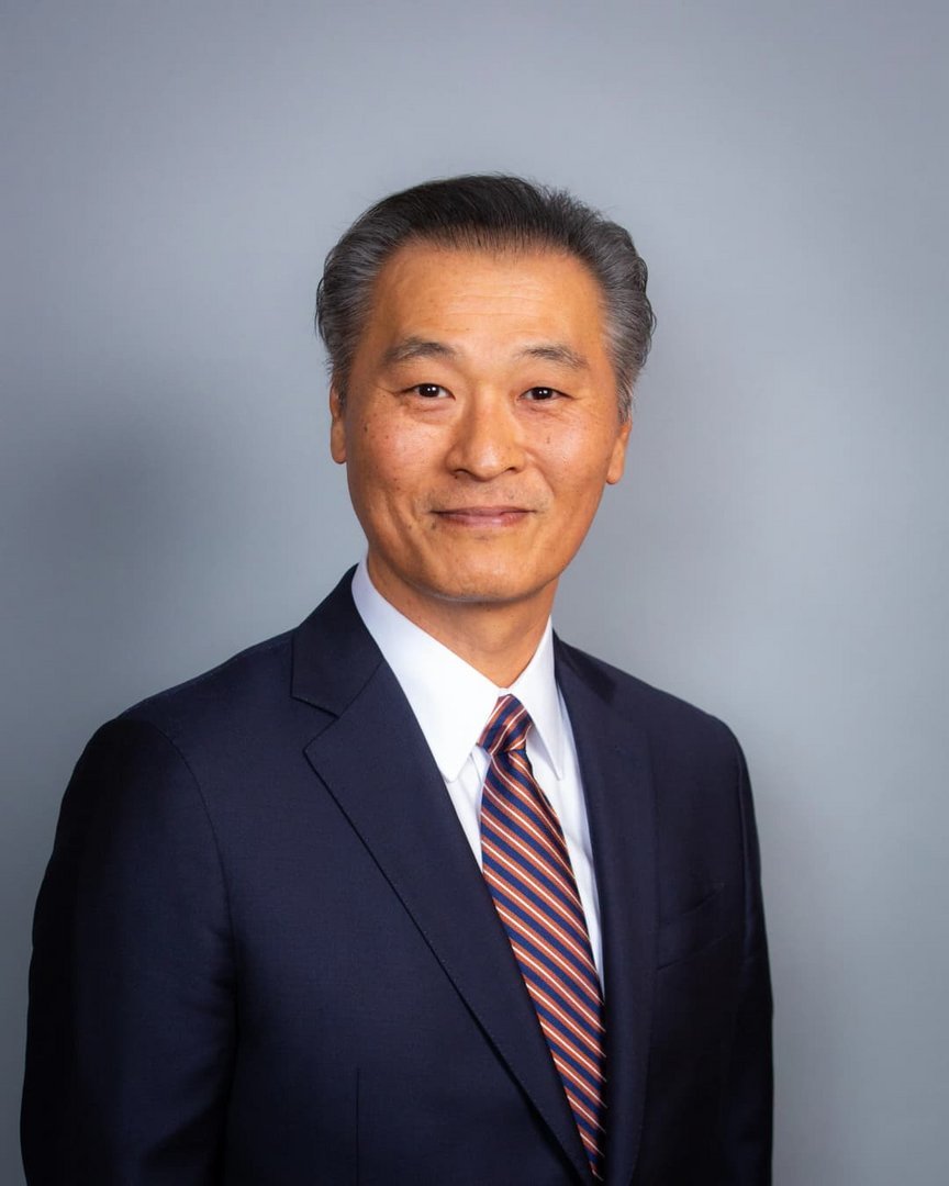 Middle-aged East Asian man in navy blue suit and red-blue striped tie smiling against neutral gray studio background