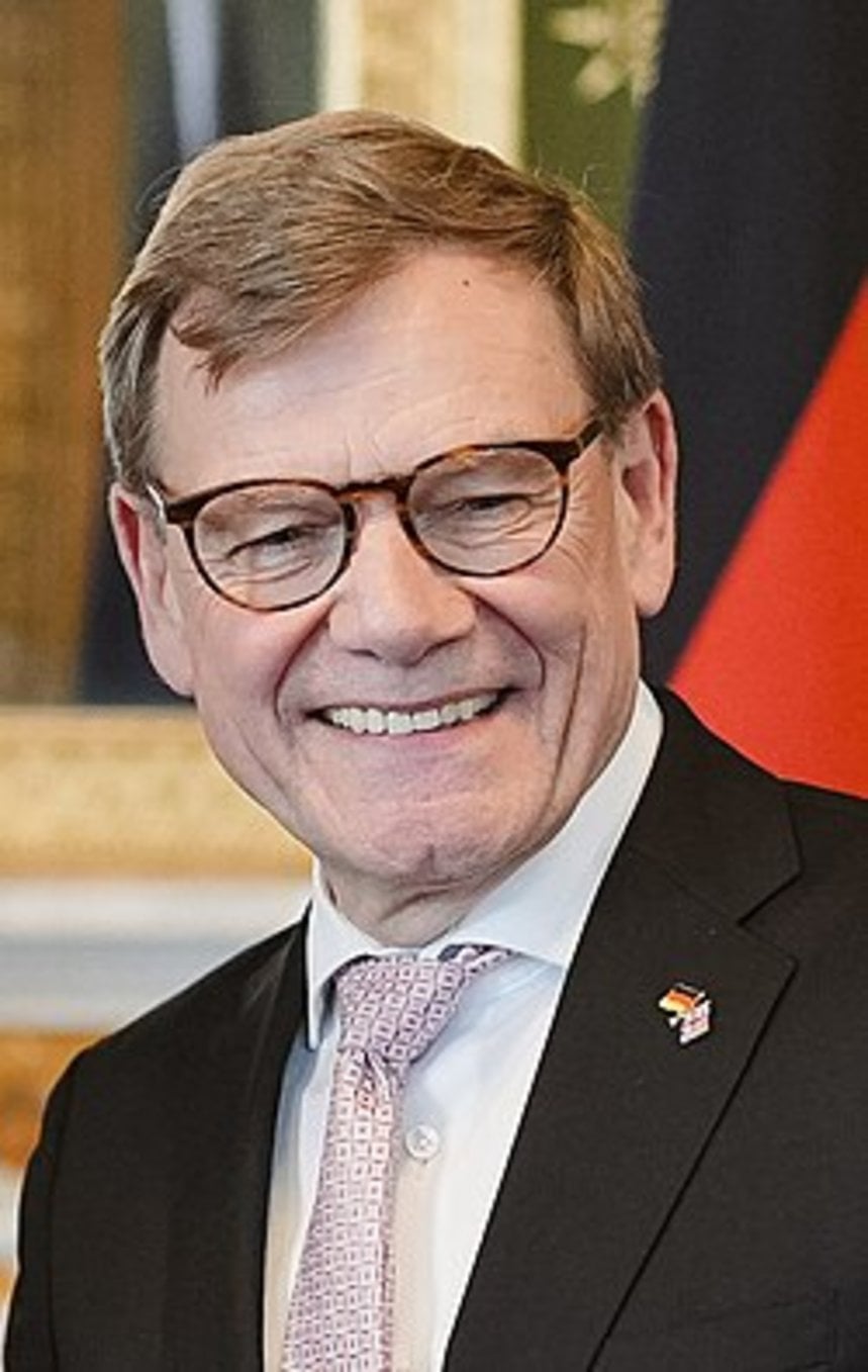 Smiling middle-aged man with round tortoiseshell glasses, gray-blonde hair, dark suit, patterned tie, and German-UK flag lape