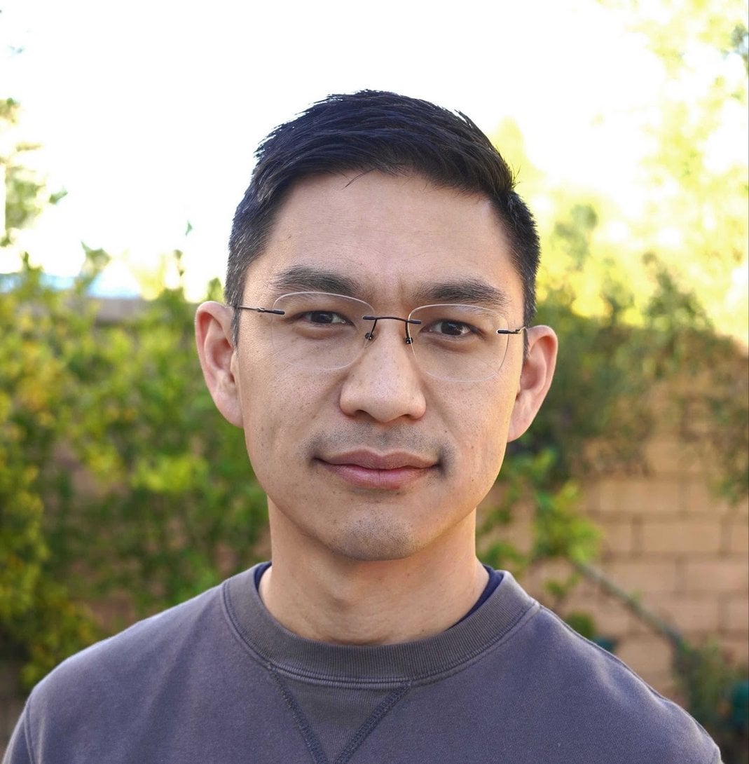 Young Asian man with short black hair wearing rimless glasses and a gray crewneck sweatshirt, standing outdoors near green fo