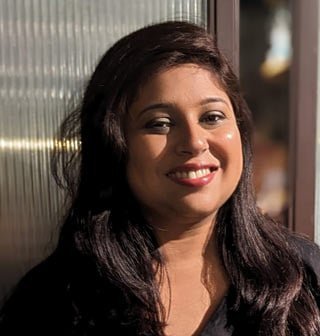 A cheerful South Asian woman with long dark wavy hair and red lipstick smiling warmly in soft ambient evening lighting