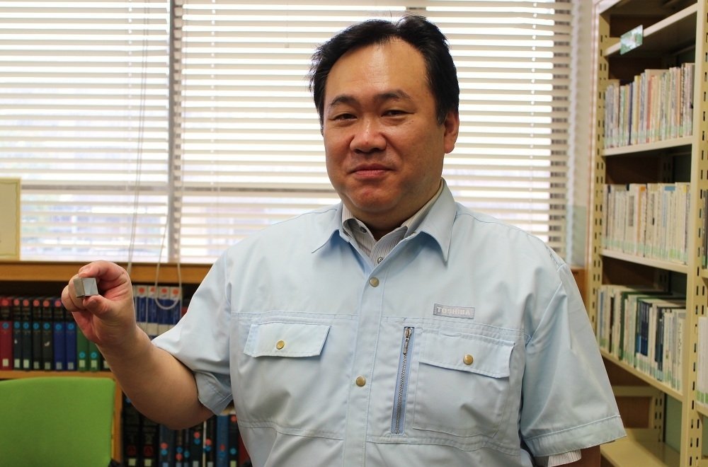 Toshiba employee in light blue uniform shirt holding a small white electronic component in a library with bookshelves