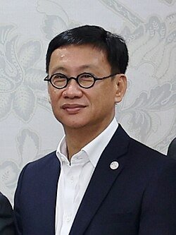 Middle-aged Asian man wearing round black-framed glasses and a navy blue suit with white shirt at a formal event