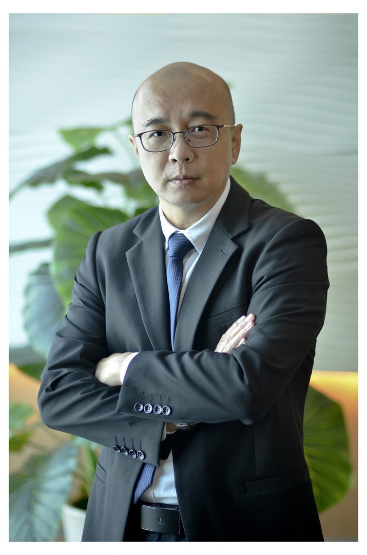 Middle-aged Asian male executive in dark charcoal suit and blue tie, arms crossed, wearing glasses, posed before indoor plant