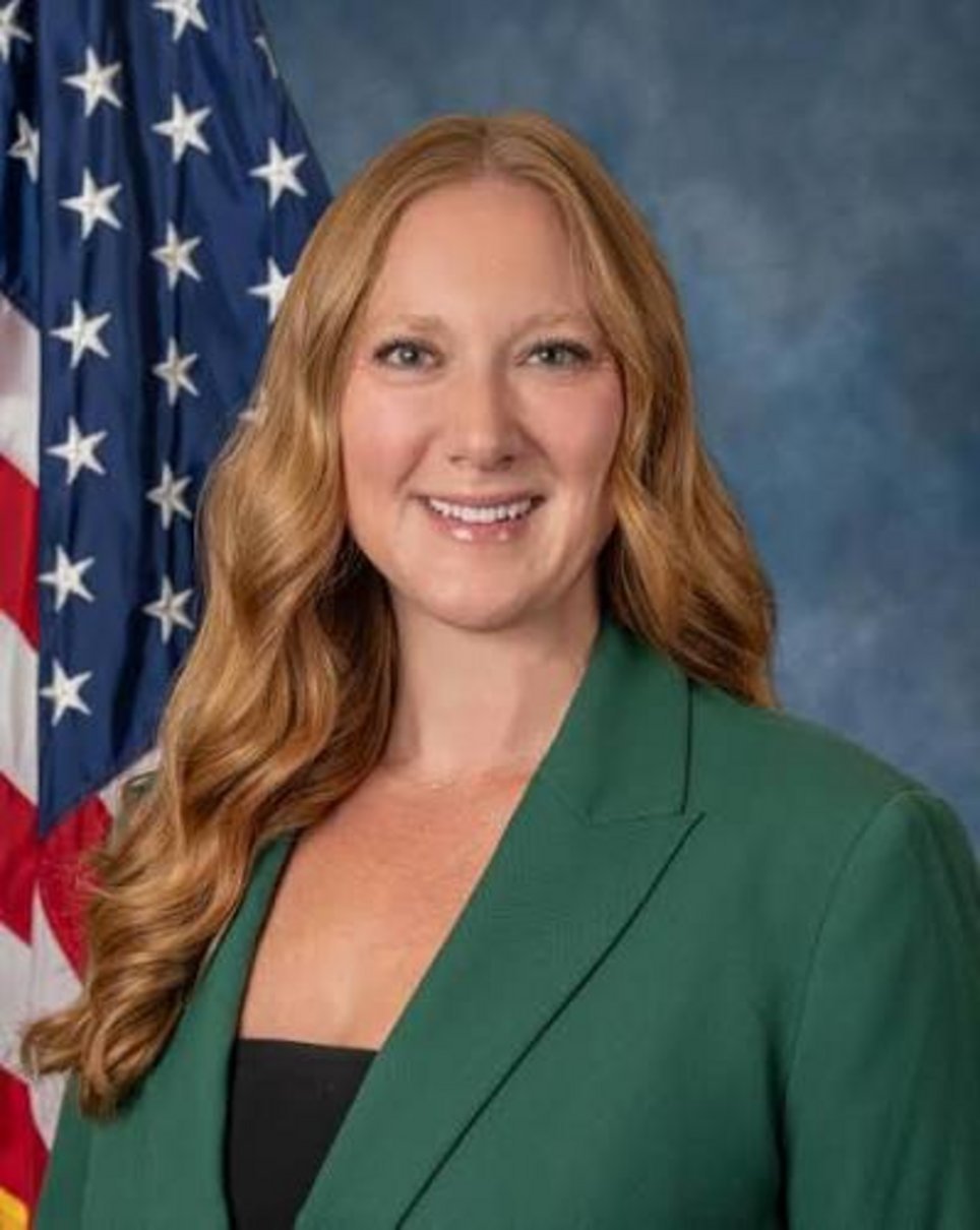 Woman with auburn wavy hair wearing a green blazer and black top, smiling in front of a United States flag on a blue backdrop