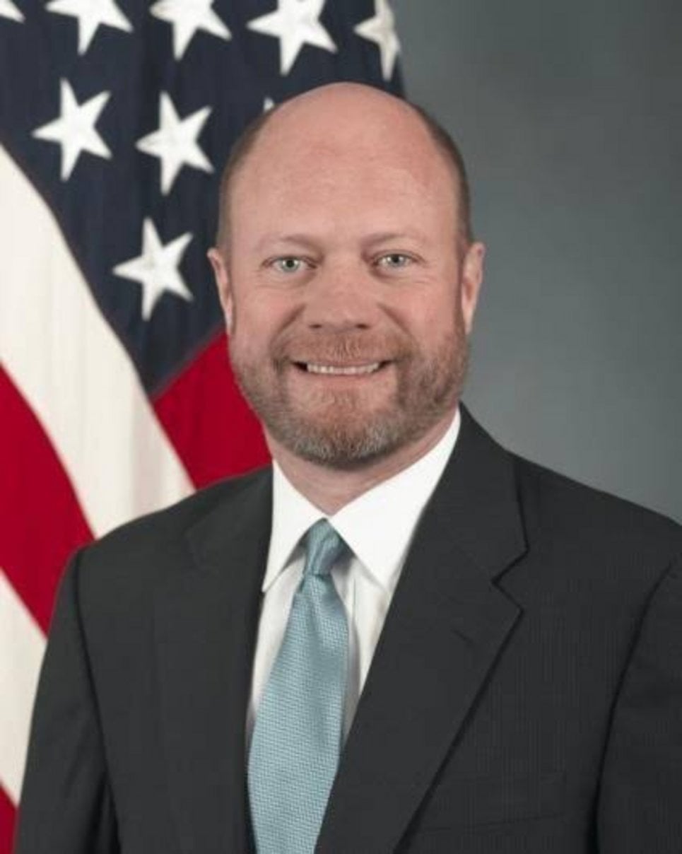 Bald man with beard in dark suit and light blue tie smiling in front of American flag during official US government portrait