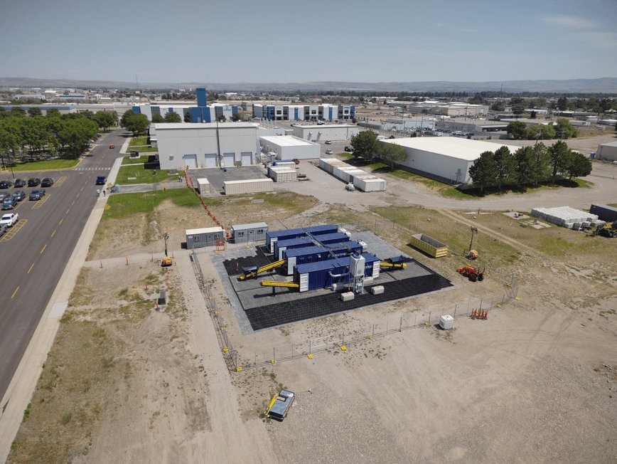 Blue containerized battery energy storage system inside security fencing at industrial park with warehouse buildings and arid