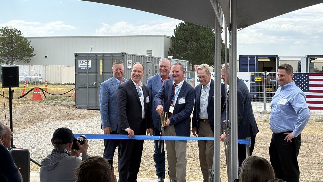 Group of business executives in suits cutting blue ribbon at outdoor industrial facility groundbreaking ceremony with America