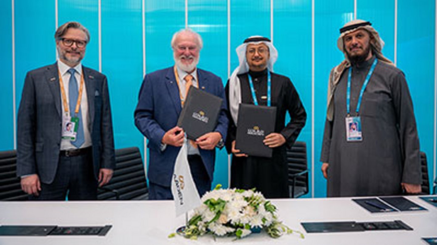 Four men in formal attire, two in Western suits and two in traditional Saudi thobes, holding signed agreement folders at a co