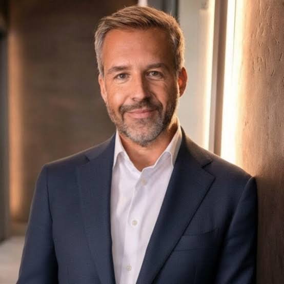 Middle-aged man with salt-and-pepper hair and short beard wearing navy blazer and white shirt, leaning against wooden wall in