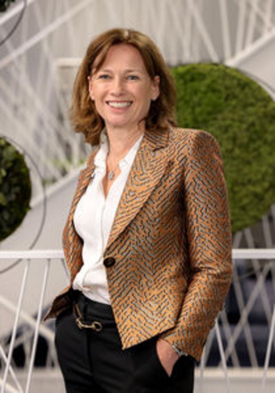 Smiling middle-aged woman with shoulder-length auburn hair wearing an orange patterned blazer and white blouse, standing outd