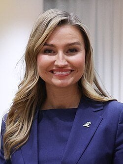 Young blonde woman with long wavy hair wearing a navy blue blazer with a gold lapel pin, smiling warmly indoors