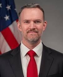 Professional man with short gray beard wearing dark suit and red tie posed before American flag in official government portra