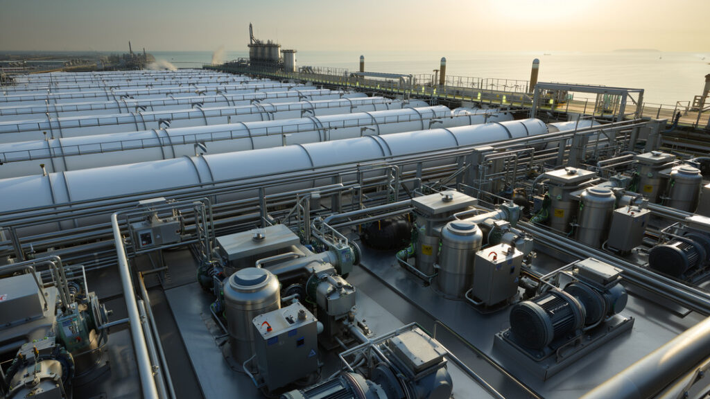 Photorealistic wide-angle documentary view of **water desalination plants** at golden hour, showing stainless-steel high-pressure pump skids and dense pipework in the foreground, parallel rows of white RO membrane vessels midground, and the ocean intake/outfall with a hazy industrial coastline in the background.