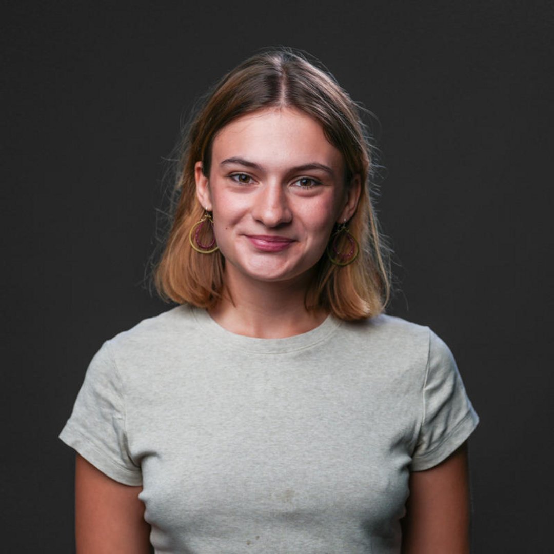 Young woman with shoulder-length blonde hair and gold hoop earrings wearing a light gray t-shirt, smiling softly against dark