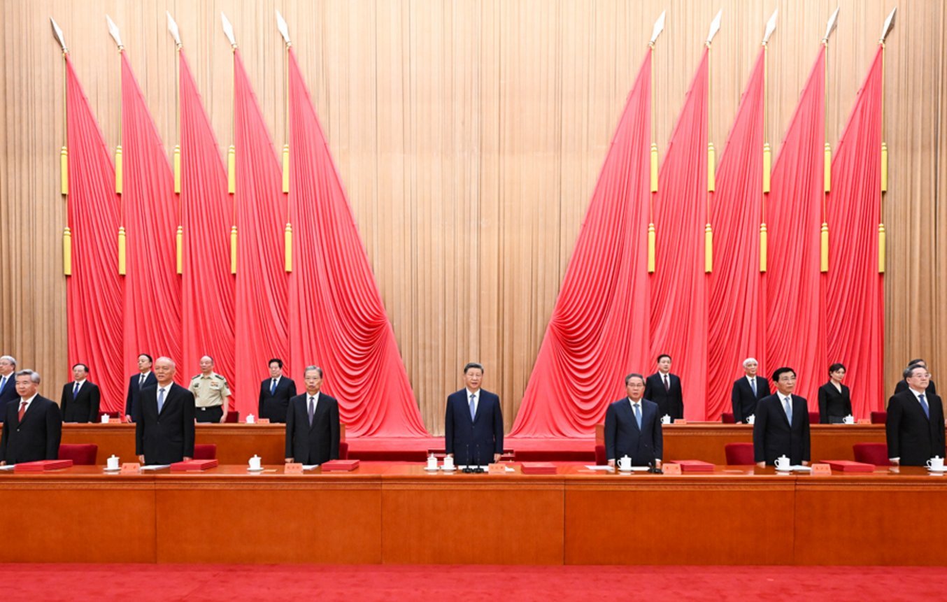 Senior Chinese government officials in dark suits standing at podium before large red ceremonial flags at formal state event