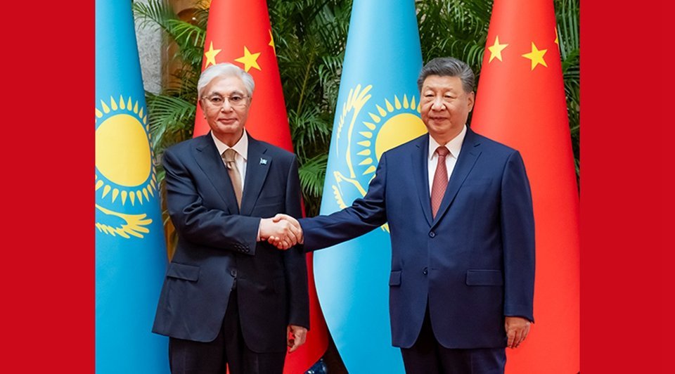Two officials in dark suits shaking hands before Kazakhstan light-blue and Chinese red national flags at formal bilateral sum